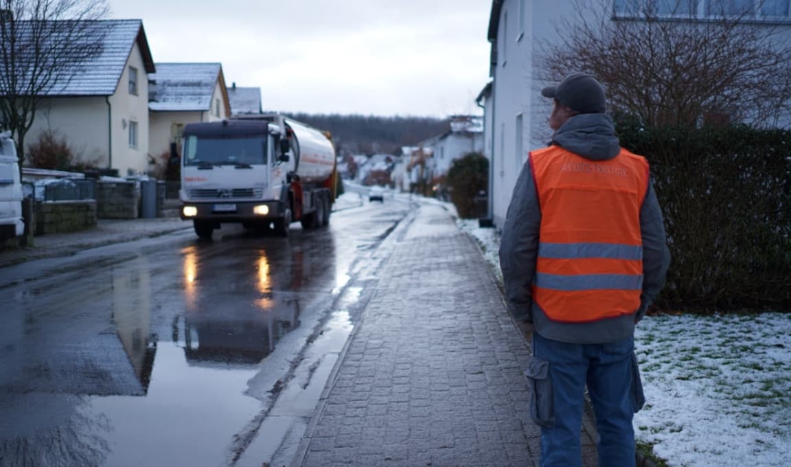Człowiek w kamizelce odblaskowej stoi na chodniku, drogą przejeżdża pojazd asenizacyjny